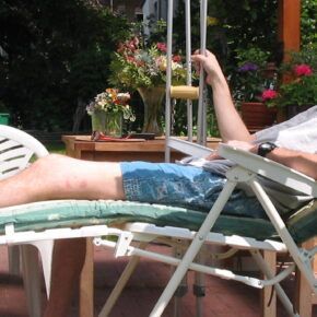 Verfasser im Liegestuhl auf der Terrasse, Krcken neben sich. Er trägt kurze Hosen, ein T-Shirt und sein linker Fuß ist verbunden. Die Sonne scheint auf den Garten im Hintergrund