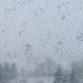 Fensterscheibe mit Schneeflocken und im Hintergrund verschwommen Felder und Bäume