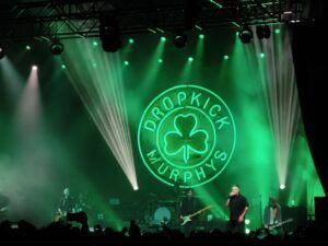 Die Dropkick Murphys auf der Bühne vor grüner Beleuchtung, einer großen grünen Neon-Vignette mit dem Bandnamen und einem Kleeblatt in der Mitte