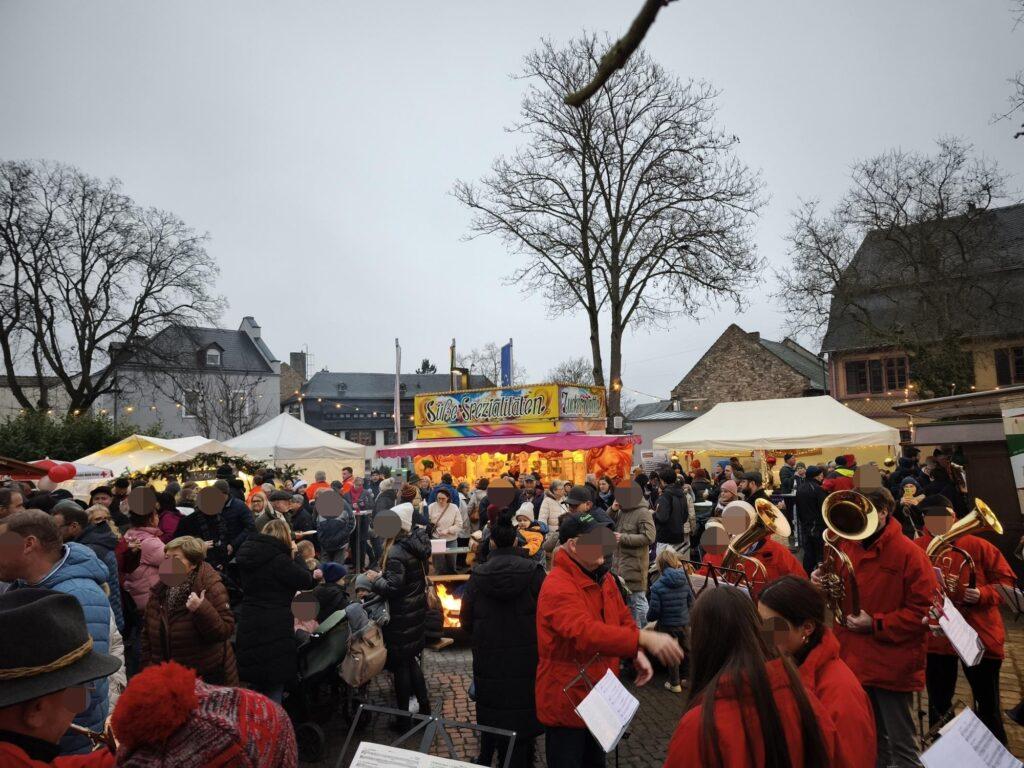 Weihnachtsmarkt Oestrich-Winkel, Foto vom Areal aus Eingangssicht mit Bläsergruppe, Menschen, Zelten und Süßigkeitenstand, im Hintergrund der Ort.