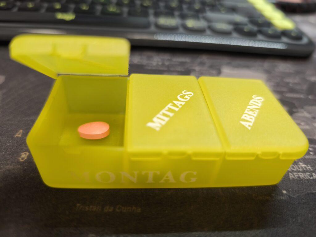 Eine Tablettenschachtel aus gelbem Kunststoff mit der Aufschrift 'Montag' und Einteilungen in Morgens, Mittags, Abends. Das 'Morgens'-Fach ist geöffnet und eine orangefarbene Tablette ist sichtbar.