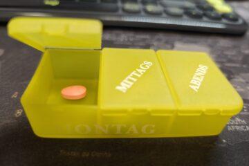 Eine Tablettenschachtel aus gelbem Kunststoff mit der Aufschrift 'Montag' und Einteilungen in Morgens, Mittags, Abends. Das 'Morgens'-Fach ist geöffnet und eine orangefarbene Tablette ist sichtbar.