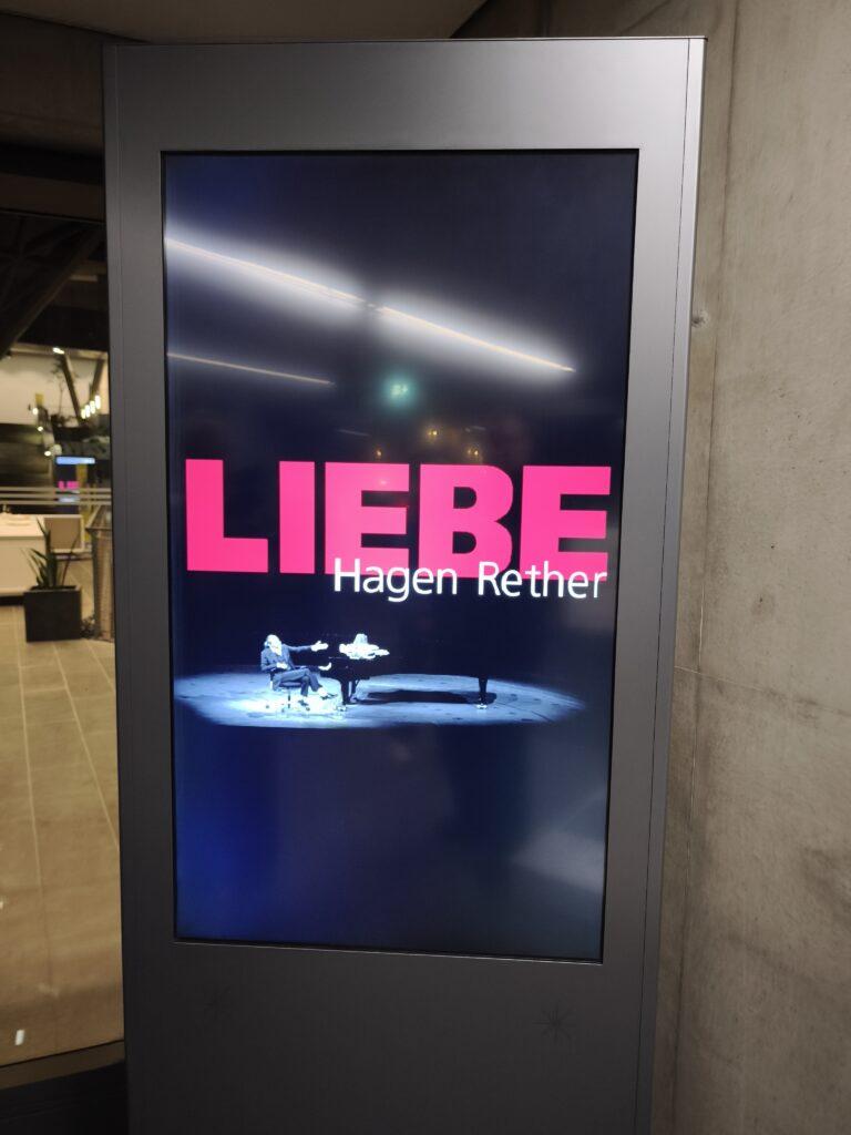 Digitaler Schaukasten mit der Ankündigung 'Liebe' und 'Hagen Rether', im Hintergrund besagter Herr mit einem mit Bananen belegtem schwarzen Konzertflügel