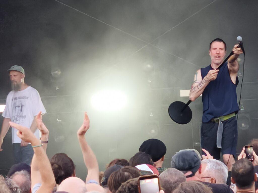Sleaford Mods auf der Bühne, Köpf des Publikums im Vordergrund, Jason Williamson hält das Mikro in die Menge